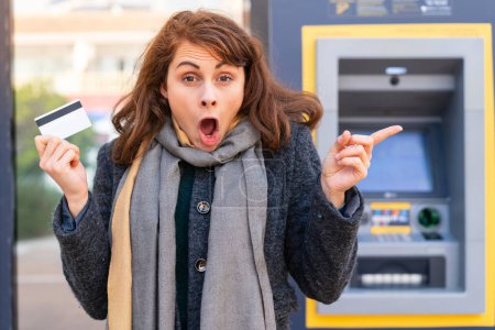 Téléchargez les photos : Femme brune tenant une carte de crédit à l'extérieur surpris et pointant du doigt vers le côté - en image libre de droit