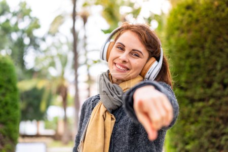 Téléchargez les photos : Brunette femme écoute de la musique avec des écouteurs à l'extérieur pointe du doigt vers vous avec une expression confiante - en image libre de droit
