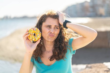 Photo for Young sport woman holding a donut at outdoors having doubts and with confuse face expression - Royalty Free Image