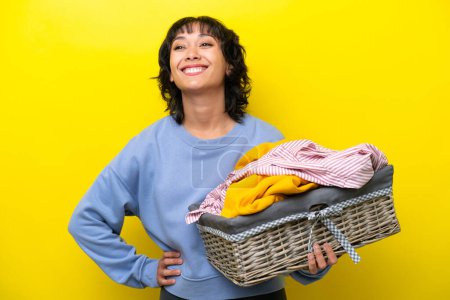 Téléchargez les photos : Jeune Argentin tenant un panier à vêtements isolé sur fond jaune posant les bras à la hanche et souriant - en image libre de droit