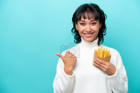 Foto de Joven argentina sosteniendo chips fritos aislados sobre fondo azul apuntando hacia un lado para presentar un producto - Imagen libre de derechos