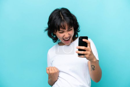 Téléchargez les photos : Jeune femme argentine isolée sur fond bleu à l'aide d'un téléphone portable et faisant un geste de victoire - en image libre de droit
