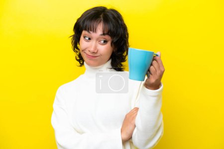 Foto de Joven argentina sosteniendo taza de café aislada sobre fondo amarillo con expresión triste - Imagen libre de derechos