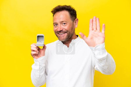 Téléchargez les photos : Homme du Moyen Age tenant une bague de fiançailles isolée sur fond jaune saluant à la main avec une expression heureuse - en image libre de droit