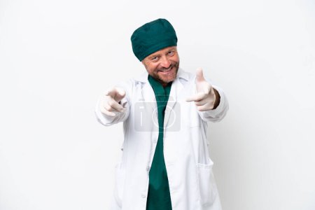 Téléchargez les photos : Chirurgien d'âge moyen en uniforme vert isolé sur fond blanc pointe du doigt vers vous tout en souriant - en image libre de droit