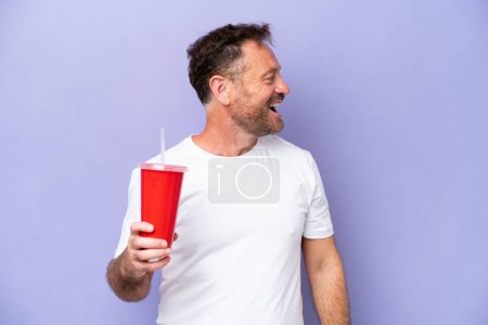 Foto de Hombre caucásico de mediana edad sosteniendo soda aislada sobre fondo púrpura riendo en posición lateral - Imagen libre de derechos