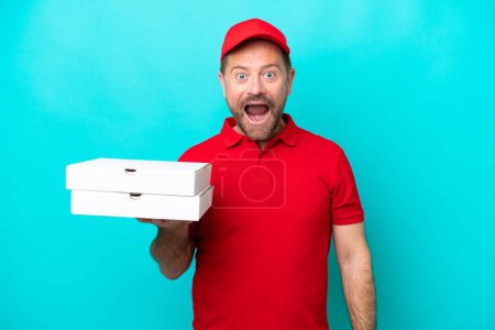 Téléchargez les photos : Livreur de pizza avec uniforme de travail ramasser des boîtes de pizza isolées sur fond bleu avec expression faciale surprise - en image libre de droit