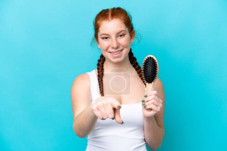 Téléchargez les photos : Jeune femme caucasienne rougeâtre avec peigne à cheveux isolé sur fond bleu pointe doigt vers vous avec une expression confiante - en image libre de droit