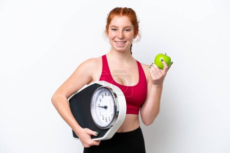 Téléchargez les photos : Young reddish woman isolated on white background with weighing machine and with an apple - en image libre de droit