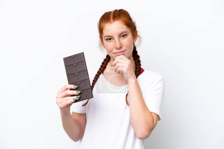 Téléchargez les photos : Jeune femme rougeâtre isolée sur fond blanc prenant un comprimé de chocolat et ayant des doutes - en image libre de droit