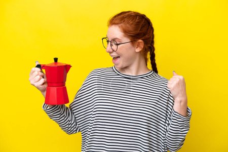 Téléchargez les photos : Jeune femme rougeâtre tenant une cafetière isolée sur fond jaune célébrant une victoire - en image libre de droit