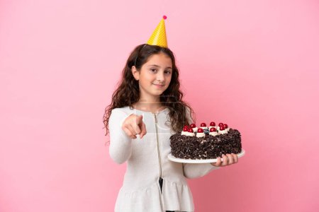 Téléchargez les photos : Petite fille caucasienne tenant gâteau d'anniversaire isolé sur fond rose pointe doigt à vous avec une expression confiante - en image libre de droit