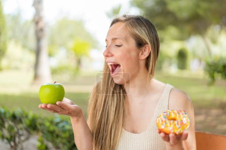 Photo for Young blonde woman at outdoors holding apple and donut with happy expression - Royalty Free Image