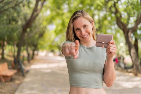 Téléchargez les photos : Jeune femme blonde tenant un portefeuille à l'extérieur pointe du doigt vers vous avec une expression confiante - en image libre de droit