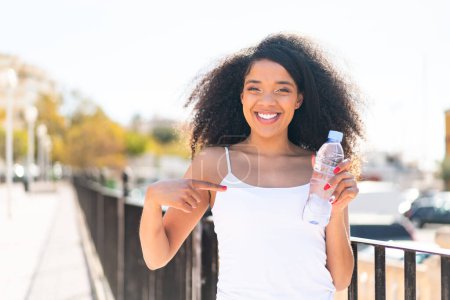 Téléchargez les photos : Jeune femme afro-américaine avec une bouteille d'eau à l'extérieur avec une expression faciale surprise - en image libre de droit