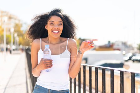 Foto de Joven mujer afroamericana con una botella de agua al aire libre sorprendida y señalando con el dedo hacia un lado - Imagen libre de derechos