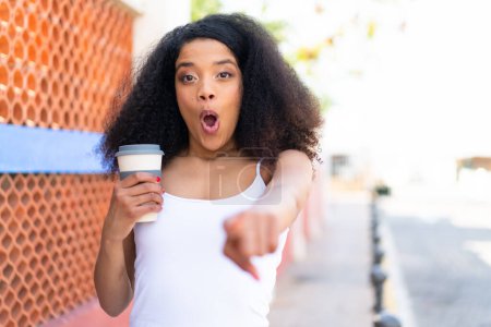 Photo for Young African American woman holding a take away coffee at outdoors surprised and pointing front - Royalty Free Image
