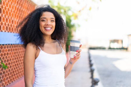 Téléchargez les photos : Jeune femme afro-américaine tenant un café à emporter à l'extérieur souriant beaucoup - en image libre de droit