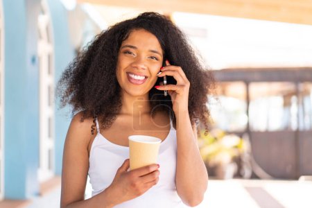 Téléchargez les photos : Jeune femme afro-américaine à l'extérieur utilisant un téléphone portable et tenant un café avec une expression heureuse - en image libre de droit