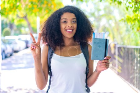 Téléchargez les photos : Jeune femme afro-américaine tenant un passeport à l'extérieur souriant et montrant signe de victoire - en image libre de droit