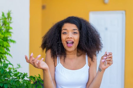 Téléchargez les photos : Jeune femme afro-américaine tenant les clés de la maison à l'extérieur avec une expression faciale choquée - en image libre de droit
