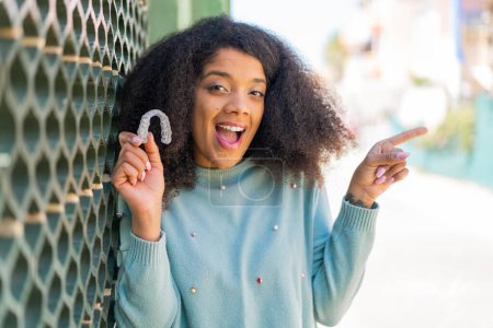 Téléchargez les photos : Jeune femme afro-américaine tenant des bretelles invisibles à l'extérieur surpris et pointant du doigt vers le côté - en image libre de droit