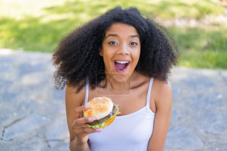 Téléchargez les photos : Jeune femme afro-américaine tenant un hamburger à l'extérieur avec une expression faciale surprise et choquée - en image libre de droit
