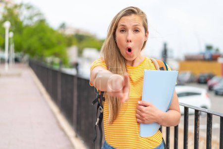 Téléchargez les photos : Jeune étudiante blonde femme à l'extérieur surpris et pointant devant - en image libre de droit