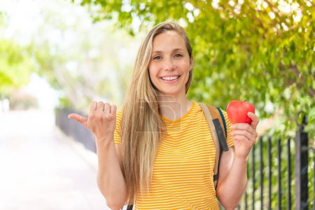 Téléchargez les photos : Jeune femme blonde avec une pomme à l'extérieur pointant vers le côté pour présenter un produit - en image libre de droit
