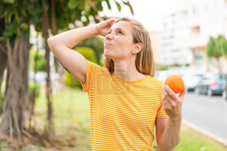 Téléchargez les photos : Jeune femme blonde tenant une orange à l'extérieur ayant des doutes et avec une expression du visage confuse - en image libre de droit