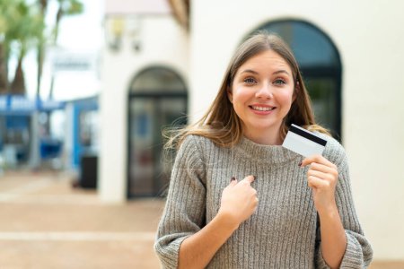 Foto de Joven mujer rubia bonita sosteniendo una tarjeta de crédito al aire libre con expresión facial sorpresa - Imagen libre de derechos