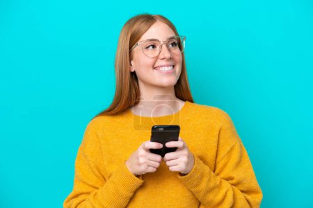 Téléchargez les photos : Jeune rousse femme isolée sur fond bleu en utilisant un téléphone mobile et en levant les yeux - en image libre de droit