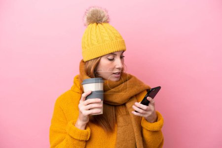 Photo for Young redhead woman wearing winter jacket isolated on pink background holding coffee to take away and a mobile - Royalty Free Image