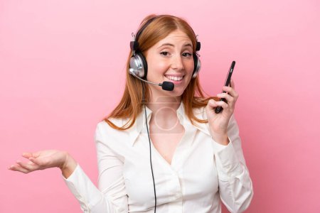 Photo for Telemarketer redhead woman working with a headset isolated on pink background keeping a conversation with the mobile phone with someone - Royalty Free Image