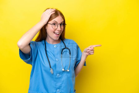 Foto de Joven enfermera pelirroja aislada sobre fondo amarillo sorprendida y señalando con el dedo hacia un lado - Imagen libre de derechos