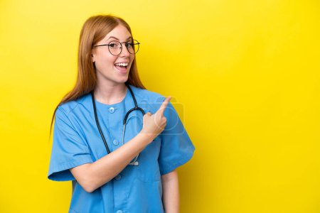 Foto de Joven enfermera pelirroja aislada sobre fondo amarillo sorprendida y señalando lado - Imagen libre de derechos