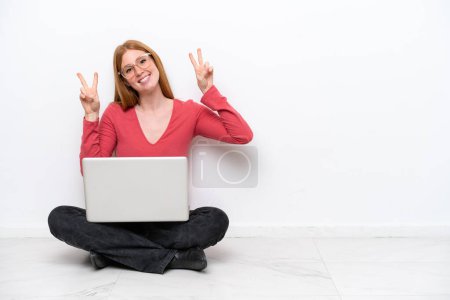 Photo for Young redhead woman with a laptop sitting on the floor isolated on white background showing victory sign with both hands - Royalty Free Image