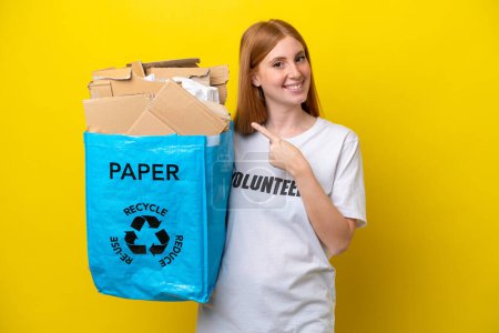 Foto de Joven pelirroja sosteniendo una bolsa de reciclaje llena de papel para reciclar aislada sobre fondo amarillo apuntando hacia un lado para presentar un producto - Imagen libre de derechos