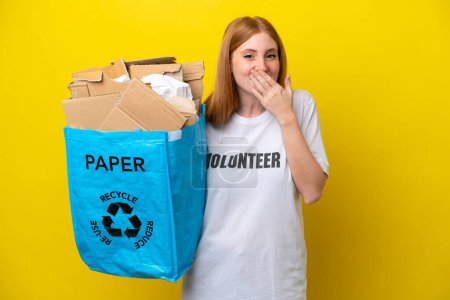 Téléchargez les photos : Jeune rousse femme tenant un sac de recyclage plein de papier à recycler isolé sur fond jaune heureux et souriant couvrant la bouche avec la main - en image libre de droit