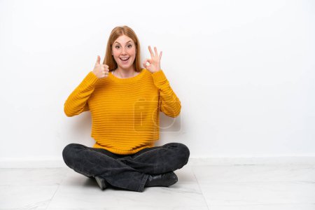 Téléchargez les photos : Jeune rousse femme assise sur le sol isolé sur fond blanc montrant signe ok et pouce geste vers le haut - en image libre de droit