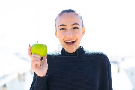 Foto de Chica joven marroquí con una manzana al aire libre con sorpresa y expresión facial conmocionada - Imagen libre de derechos