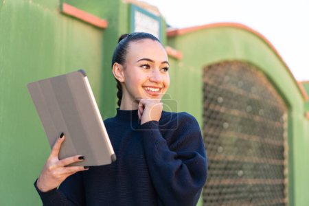 Téléchargez les photos : Jeune fille marocaine tenant une tablette à l'extérieur pensant une idée et regardant côté - en image libre de droit