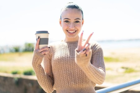 Photo for Young moroccan girl holding a take away coffee at outdoors smiling and showing victory sign - Royalty Free Image