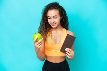 Photo for Young Arab woman isolated on blue background taking a chocolate tablet in one hand and an apple in the other - Royalty Free Image