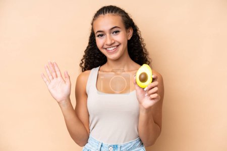 Photo for Young woman holding an avocado isolated on beige background saluting with hand with happy expression - Royalty Free Image