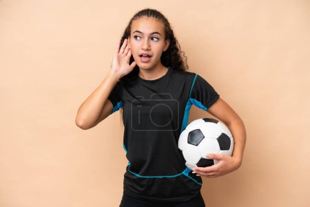 Photo for Young football player woman isolated on beige background listening to something by putting hand on the ear - Royalty Free Image