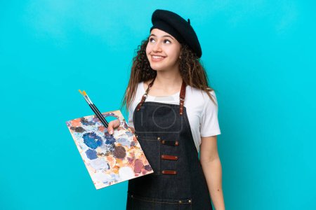 Téléchargez les photos : Jeune artiste arabe tenant une palette isolée sur fond bleu penser une idée tout en levant les yeux - en image libre de droit