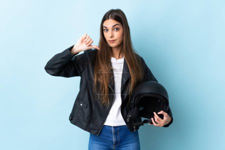 Foto de Mujer caucásica joven sosteniendo un casco de motocicleta aislado sobre fondo azul mostrando el pulgar hacia abajo con expresión negativa - Imagen libre de derechos