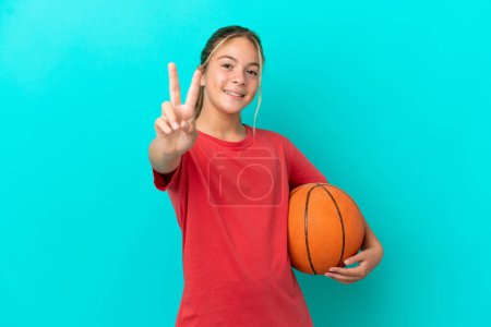 Téléchargez les photos : Petite fille caucasienne jouant au basket isolé sur fond bleu souriant et montrant le signe de victoire - en image libre de droit