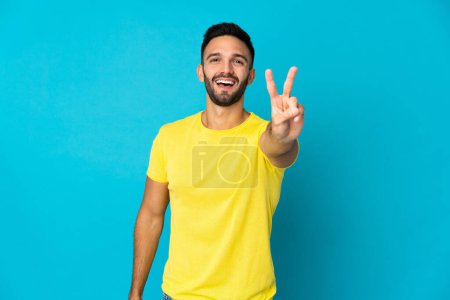 Téléchargez les photos : Jeune homme caucasien isolé sur fond bleu souriant et montrant signe de victoire - en image libre de droit
